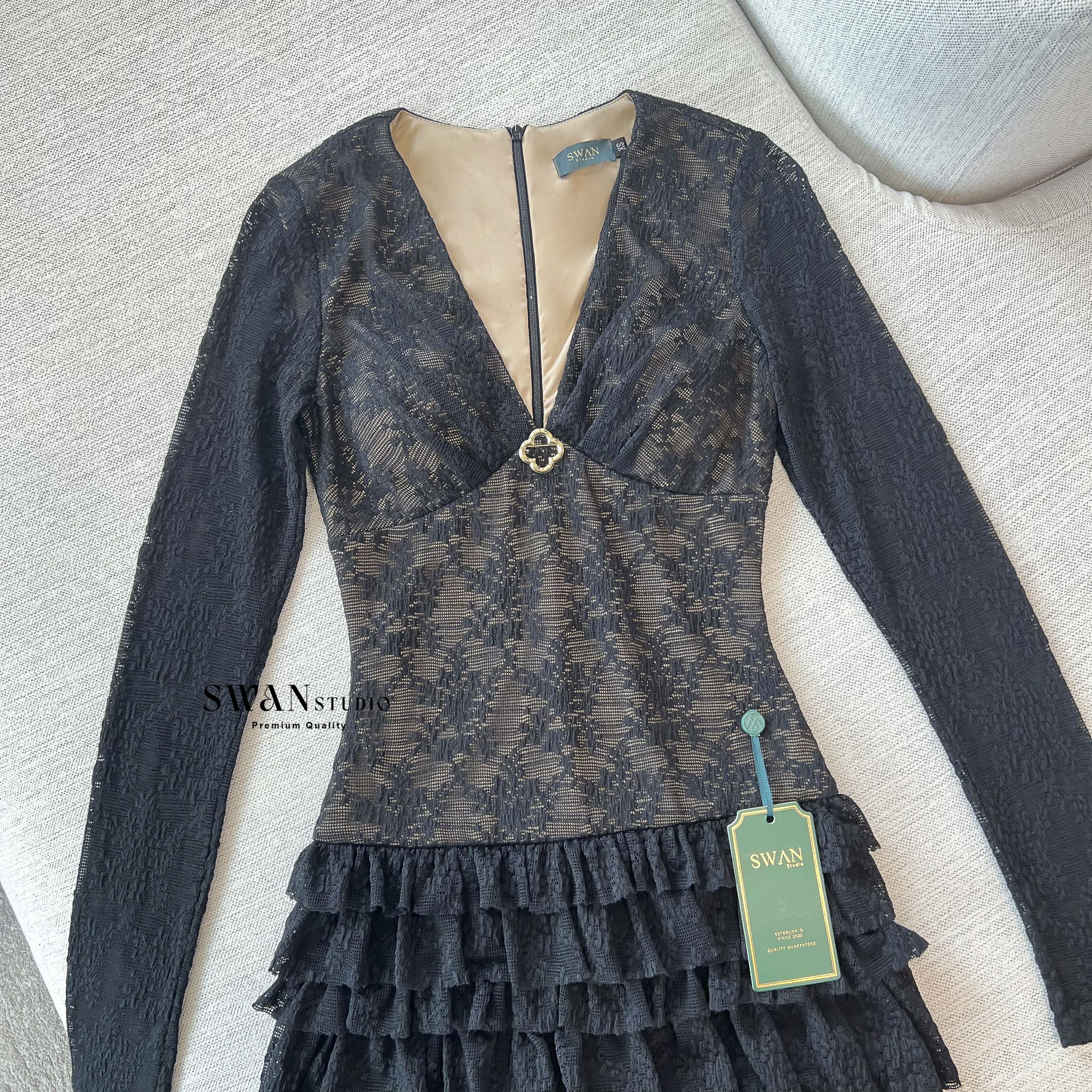 เดรสลูกไม้สีดำแขนยาวซีทรูซ้อนซับสีเนื้อ ดีไซน์เข้ารูปช่วงตัวคอวีลึกแต่งเข็มกลัดสีทองช่วงอกเพิ่มความหรูหราตัดต่อกระโปรงสั้นบานระบายเลเยอร์ชั้นๆด้วยผ้าลูกไม้ดูพริ้วหวานแฝงความเซ็กซี่ลงตัวเหมาะกับลุคดินเนอร์หรือออกงานกลางคืนที่ต้องการความโดดเด่นและดูแพง